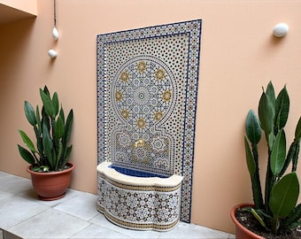 May include: A decorative mosaic fountain with a detailed geometric design in blue, gold, and white. The fountain has a curved basin and is set against a peach-colored wall, with potted plants on either side.