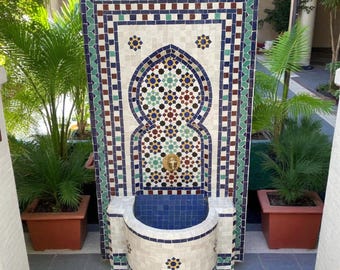 May include: A decorative outdoor fountain with a mosaic tile design. The fountain features a central arched niche with intricate patterns in blue, green, brown, and white tiles. A brass faucet is centered above a blue tiled basin. Potted plants frame the fountain.