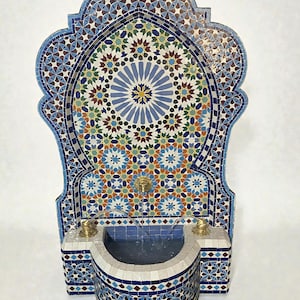 May include: A decorative mosaic fountain with a multi-colored geometric pattern. The fountain features a central basin and a curved top with a detailed mosaic design. The fountain has brass fixtures and is approximately 48 inches tall.