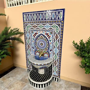 May include: A decorative outdoor fountain with a mosaic tile design. The fountain features a basin with a gold-colored spout and a patterned base. The tiles are in shades of blue, green, red, and white. The fountain is set against a peach-colored wall.