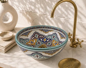 May include: A round ceramic vessel sink featuring a vibrant, multi-colored geometric pattern in shades of blue, yellow, and maroon. The sink is paired with a tall, curved brass faucet and a matching circular brass soap dish on a marble countertop.