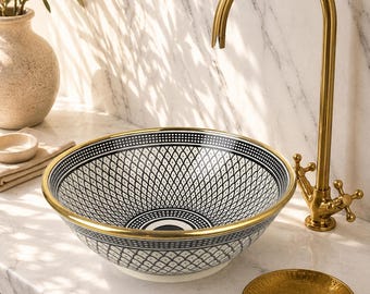 May include: A round vessel sink featuring a black and white geometric pattern with a gold rim, paired with a tall, curved gold faucet and a matching gold soap dish. The sink is set upon a white marble countertop with subtle grey veining.
