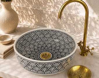 May include: A round ceramic vessel sink featuring a black and white scalloped pattern, paired with a tall, curved brass tap and a matching gold-toned drain. A small gold dish sits nearby on a light-coloured stone countertop.