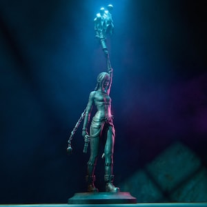 Pu&ograve; includere: Una statuetta d'argento di una donna che tiene una torcia con una luce blu e viola che le brilla addosso. La statuetta &egrave; in piedi su una base nera.