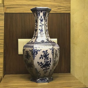 May include: A tall, hexagonal porcelain vase with a blue and white floral pattern. The vase has a wide base and a flared top, and is decorated with intricate floral designs. The vase is a decorative item.