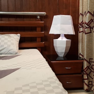 May include: A white geometric paper lamp sits on a dark nightstand with two drawers. The lamp is lit, casting a soft glow. The bedroom features a wooden headboard, patterned bedding, and a patterned curtain.