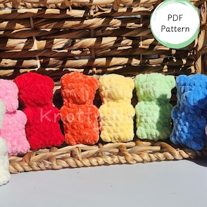 Op de afbeelding: Een rij van zes kleine, zachte, gehaakte berenfiguren in wit, roze, rood, oranje, geel, lichtgroen en blauw. Elke beer heeft een zachte, getextureerde uitstraling. Een cirkelvormig label in de rechterbovenhoek toont de tekst PDF Pattern.