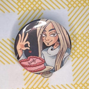 以下が含まれることがあります： 長いブロンドの髪とリップピアスをしたキャラクターが描かれた円形のピンバッジ。キャラクターは皿に乗ったハート型のピンクのケーキの上に小さなサクランボを掲げている。ケーキには筆記体でangelという文字が書かれている。
