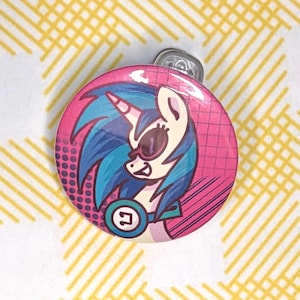May include: A circular pinback button featuring a cartoon unicorn with blue hair, wearing sunglasses and a teal headset with a white symbol. The background is a vibrant pink with a grid pattern and dark purple halftone dots on the left side.