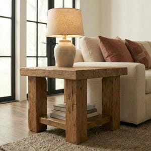 May include: A wooden side table with a lamp and books. The table has a natural wood finish and a lower shelf. The lamp has a beige shade and a cream-coloured base. A beige sofa with brown cushions is in the background.