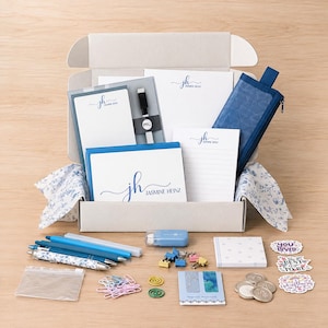 May include: A stationery gift box containing personalized notepads with the text Jasmine Heinz, a blue pencil case, pens, a blue eraser, colorful binder clips, paper clips, spiral clips, a magnetic bookmark, sticky notes, and decorative stickers.