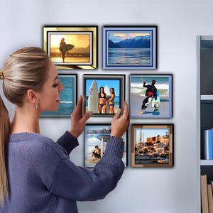 May include: A collection of framed photographs featuring various beach scenes, including surfers, a mountain range, and a cityscape. The photos are arranged in a grid pattern on a white wall.