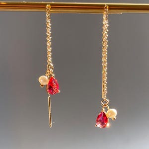 May include: A pair of gold-coloured chain earrings. Each earring features a red, teardrop-shaped gemstone and a small, white pearl. The earrings are suspended from a thin gold bar.