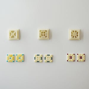 May include: Six square tiles with decorative patterns are displayed. Three are off-white with openwork designs. The other three are yellow with blue, green, and red accents. The tiles are arranged on a white surface.