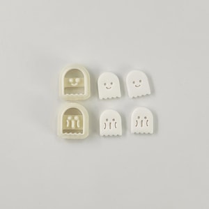 May include: A collection of white ghost-shaped cookie cutters and cut-out biscuits. The cutters have a rectangular frame with a ghost face design. The biscuits have smiling faces and a scalloped bottom edge. Ideal for Halloween baking.