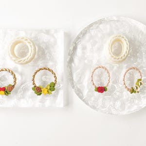 May include: Overhead view of two clear trays, one square and one round, each with a white ring and two decorative wreaths. The wreaths have gold or tan rope-like frames and are decorated with colourful floral and pinecone accents.