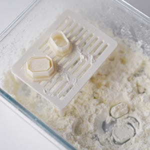 May include: A clear glass container filled with white powder, with a white plastic slotted tray holding two small, light-coloured, geometric-shaped objects. The objects appear to be moulds or cutters. The scene is brightly lit.