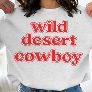 May include: A light gray sweatshirt with the words "wild desert cowboy" in red, bold, retro-style lettering. The sweatshirt is a casual, comfortable garment, perfect for everyday wear. The text is centered on the chest.