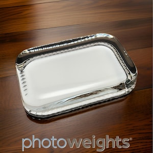 May include: A clear glass rectangular photo weight with a textured edge. The weight is designed to hold down photos or other items.