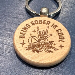May include: Wooden keychain with a peace sign, roses, and the text "Being sober is cool".