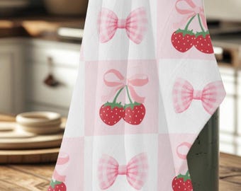 Puede incluir: Un paño de cocina a cuadros blancos y rosas con un patrón repetido de lazos rosas y fresas rojas. El paño está sobre una jarra verde en una mesa de madera.