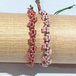 May include: Two handmade bracelets featuring pink flower patterns. The flowers are linked by green thread. The bracelets are shown on a wooden dowel against a white backdrop. These bracelets are a delicate and feminine accessory.