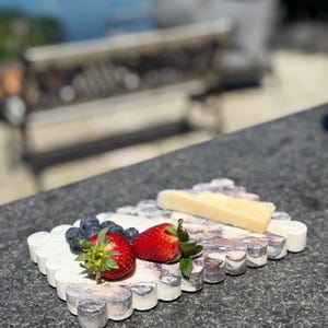 Könnte beinhalten: Ein weißes Marmor-Servierbrett mit einem gewellten Rand, auf dem eine Scheibe Käse, zwei Erdbeeren und Blaubeeren liegen. Das Brett hat ein rosa-graues Marmormuster.