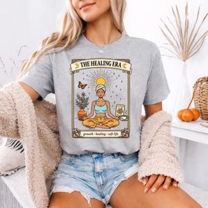 May include: A light gray t-shirt with the text "THE HEALING ERA" above a graphic of a meditating person. The graphic includes a butterfly, flowers, and the words "growth, healing, soft life".