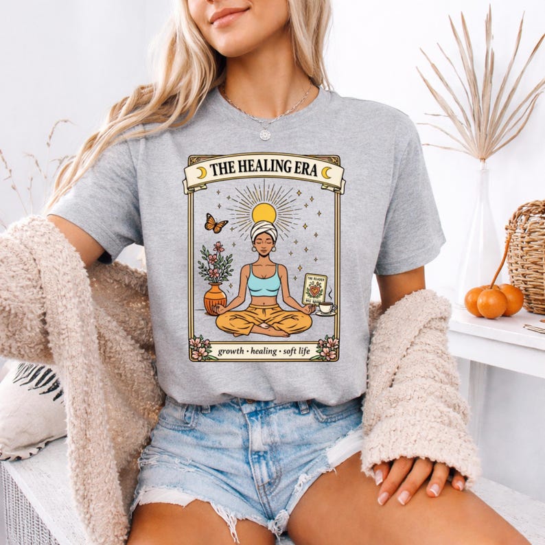 May include: A light gray t-shirt with the text "THE HEALING ERA" above a graphic of a meditating person. The graphic includes a butterfly, flowers, and the words "growth, healing, soft life".