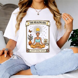 May include: White t-shirt featuring a graphic of a woman meditating with the text "THE HEALING ERA". The design includes a sun, butterfly, flowers, and the words "growth, healing, soft life".