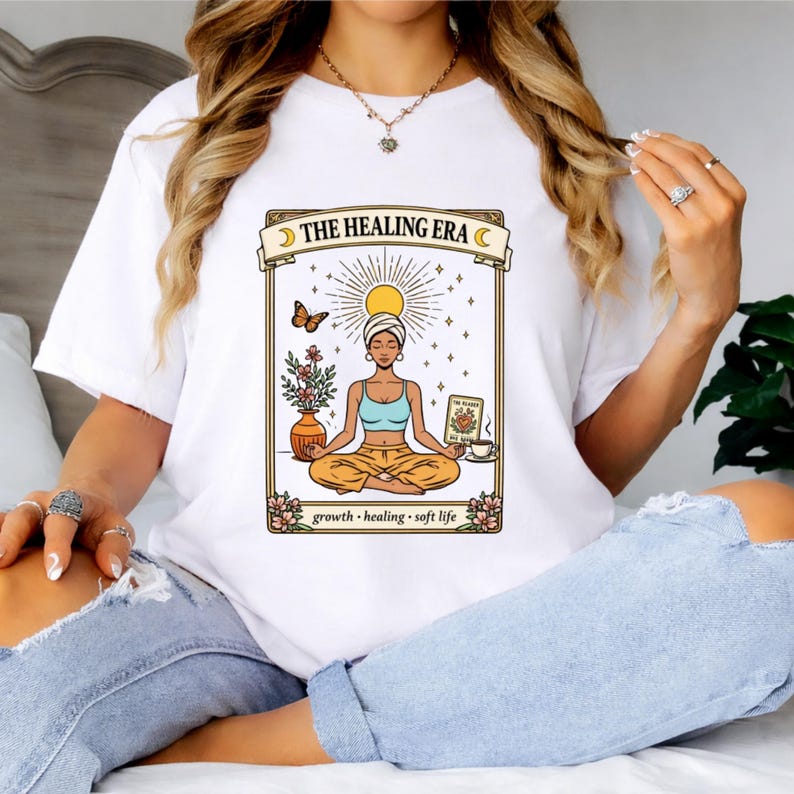 May include: White t-shirt featuring a graphic of a woman meditating with the text "THE HEALING ERA". The design includes a sun, butterfly, flowers, and the words "growth, healing, soft life".