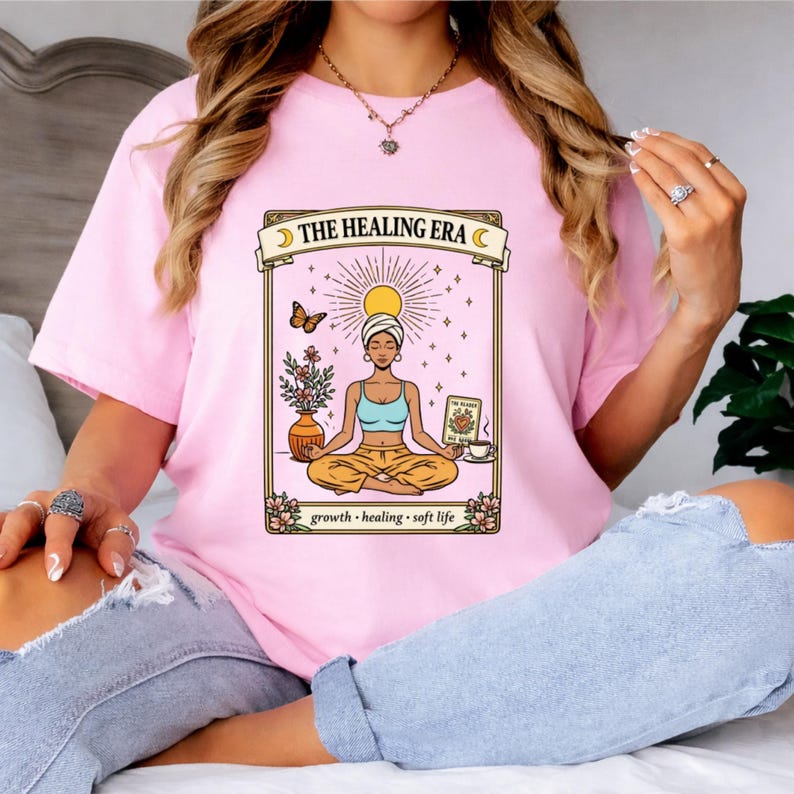 May include: A light pink t-shirt featuring a graphic of a woman meditating with the text "THE HEALING ERA". The graphic includes a sun, butterfly, flowers, and the words "growth, healing, soft life".