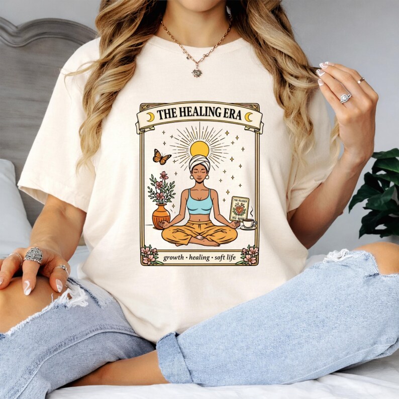 May include: Cream-colored t-shirt featuring a tarot card design with the text "THE HEALING ERA." The illustration depicts a meditating figure, a sun, a butterfly, flowers, and the words "growth, healing, soft life."