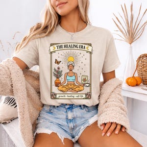May include: Beige t-shirt with a graphic featuring a meditating figure, flowers, and the text "THE HEALING ERA." The design includes a butterfly and the words "growth, healing, soft life."