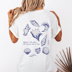 May include: White t-shirt featuring a collection of blue seashell illustrations and a wave design. The back of the shirt also includes the text "Walking in the water with my eyes fixed on days".