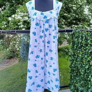 May include: A light blue nightdress with a floral pattern of blue flowers. The sleeveless nightdress is displayed on a black mannequin. The background features greenery and a glass railing.