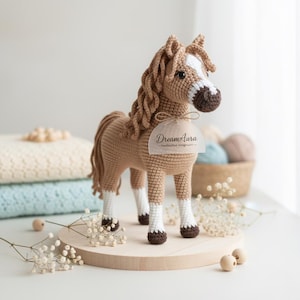 Peut inclure: Cheval en crochet fait main, aux tons marron, beige et blanc. Il présente une crinière et une queue détaillées, avec des marques blanches sur le visage et les pattes. Il est posé sur une base ronde en bois. Une étiquette avec le texte "DreamAura" est attachée.