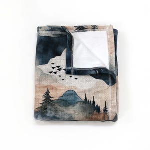May include: Folded blanket with a watercolor landscape design. The blanket features a mountain scene with trees, birds, and a color palette of blues, browns, and white. The edges are dark blue.
