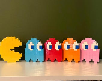 May include: A collection of five pixelated figures constructed from interlocking plastic building blocks. The set includes a yellow circular character with a wedge-shaped mouth, followed by four ghost-shaped figures in blue, red, orange, and pink.