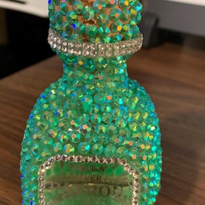 Może przedstawiać: Mała butelka tequili ozdobiona iryzującymi zielonymi kryształkami. Butelka ma srebrną opaskę i etykietę z napisem "Patron Silver". Butelka jest ozdobiona błyszczącymi kryształami, dodając odrobinę blasku.