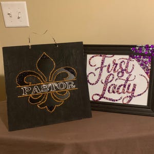 May include: Two decorative signs. One is a black square with a fleur-de-lis design and the word "PASTOR" in the center. The other is a framed sign with the words "First Lady" in a glittery script, with purple and silver embellishments.