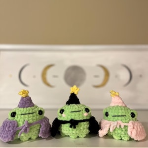 May include: Three handmade crochet frogs in a row. Each frog is green with black eyes and a small hat. One frog has a purple hat and cape, one has a black hat and cape, and one has a pink hat and cape.