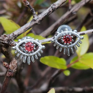 May include: A pair of silver-toned stud earrings shaped like stylized eyes, each featuring a round, faceted red gemstone at the center and radiating metal rays along the bottom edge, designed with a secure post backing for ear attachment.