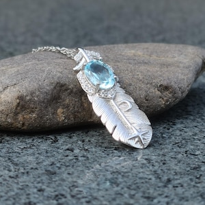 May include: A silver feather pendant necklace with a light blue oval gemstone and small clear stones. The feather design has etched details and a star at the bottom. The necklace is displayed on a gray rock.