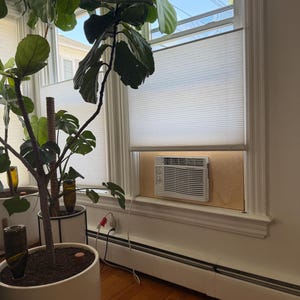 May include: A white window air conditioner unit is installed in a window opening using a light-colored wooden panel insert. The unit features a horizontal louvered front panel with control knobs on the left side and a white power cord extending down.