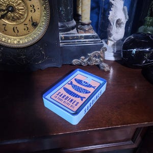 May include: A blue and pink rectangular box with the text "SARDINES IN PURE OLIVE OIL" and a graphic of three sardines. The box is sitting on a wooden surface.