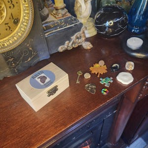 May include: A wooden box with a label that reads "Raven -  Mystical Treasures" and a variety of small trinkets, including a key, a leaf, a puzzle piece, a small eye, and a small white object.