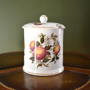 May include: A white ceramic jar with a lid, featuring a design of ripe peaches and blossoms. The jar has a gold rim and a small handle on the lid. The design includes peaches in shades of red, orange, and yellow, with green leaves and white flowers.