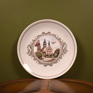 May include: A white decorative plate with a detailed image of a European town scene, including buildings and trees, framed by a gray and white ornate border. The plate has a thin gray rim and is likely a souvenir or collectible item.