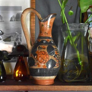 May include: A decorative ceramic pitcher with a curved handle, featuring a design of orange and black floral patterns on a cream background. The pitcher has a wide base and a narrow neck. The pitcher is displayed on a wooden shelf with other decorative items.